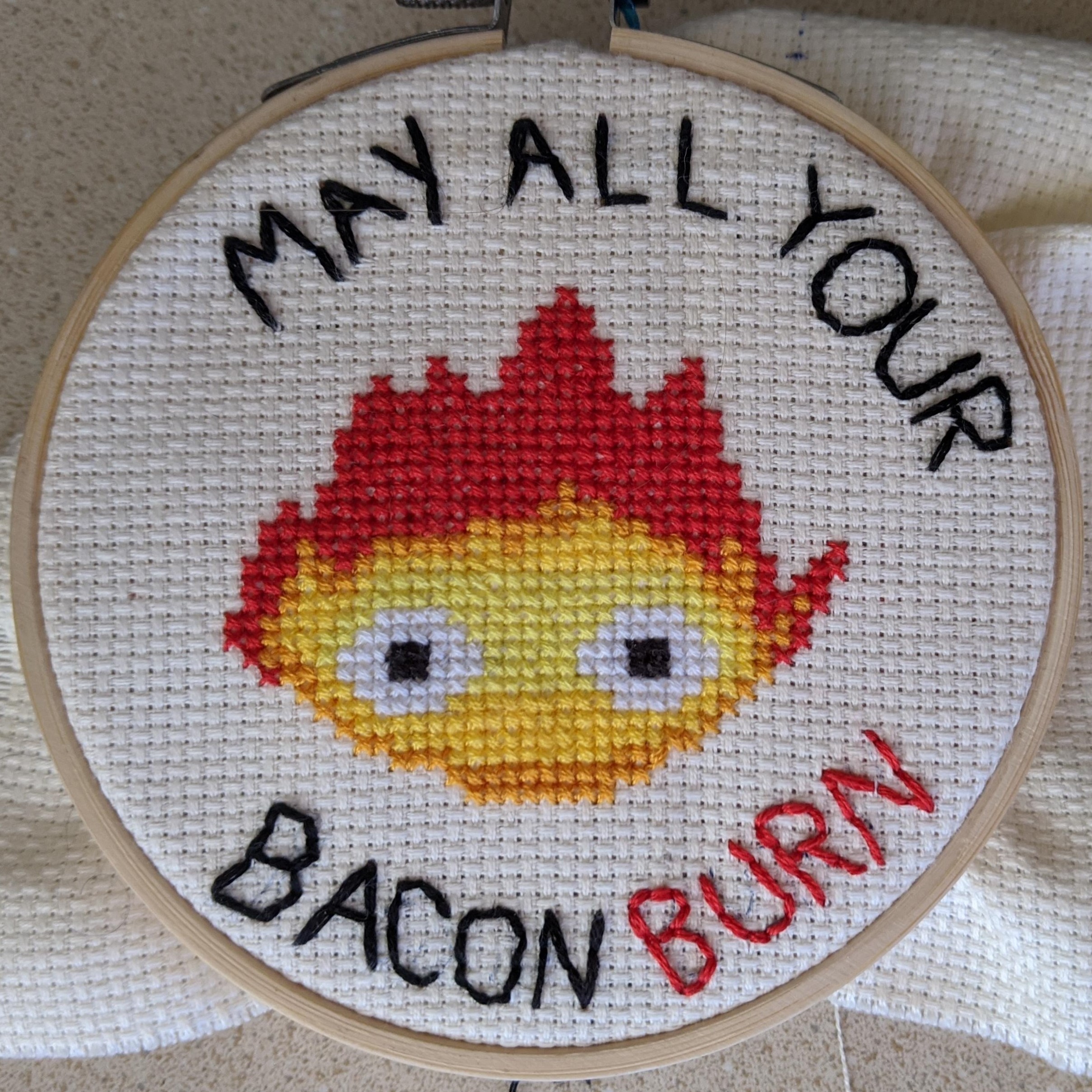 cross stitch of Calcifer from Howl's Moving Castle with the text, May all your bacon burn